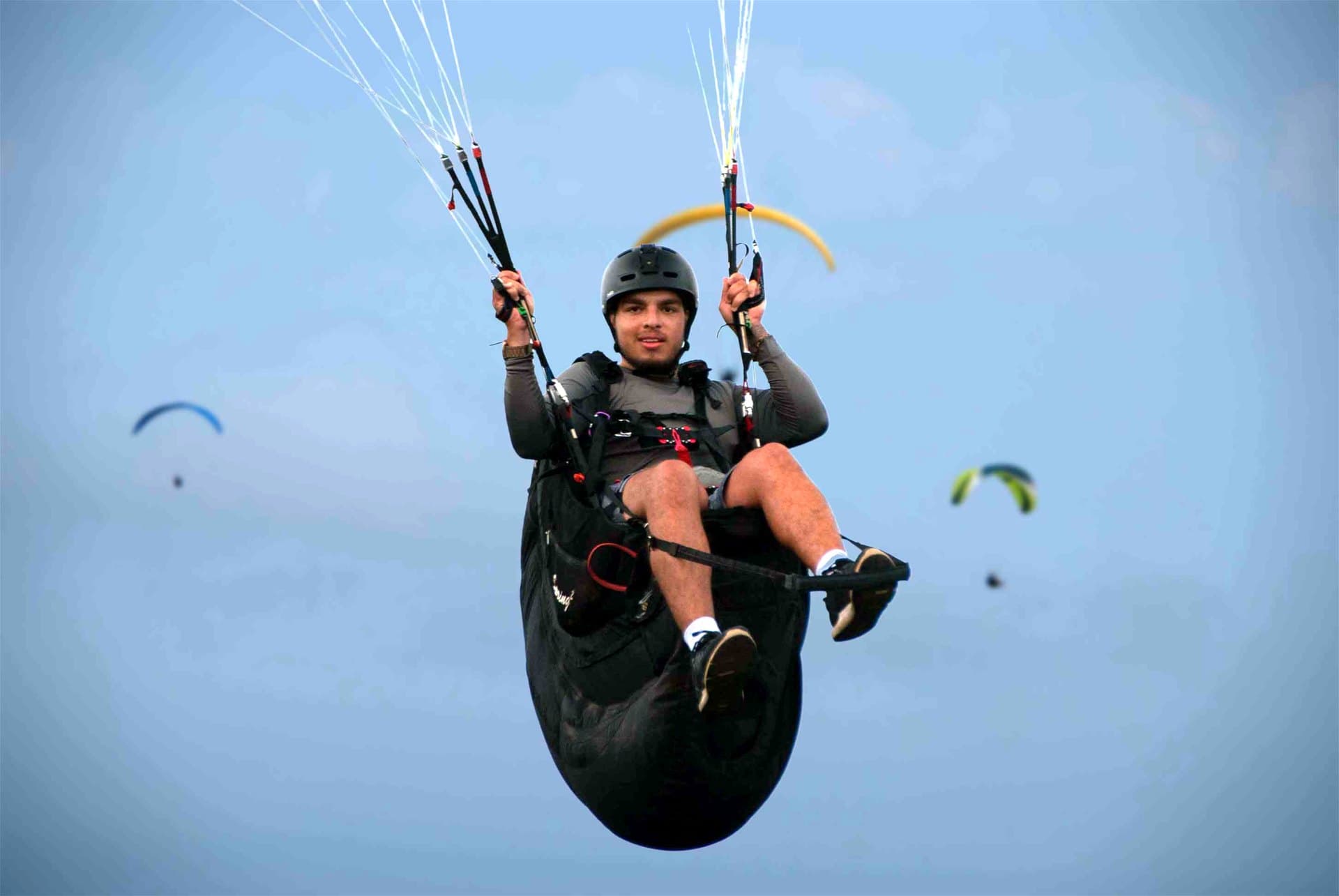 Piloto de capacete em voo, com as mãos nos comandos e outros parapentes ao fundo